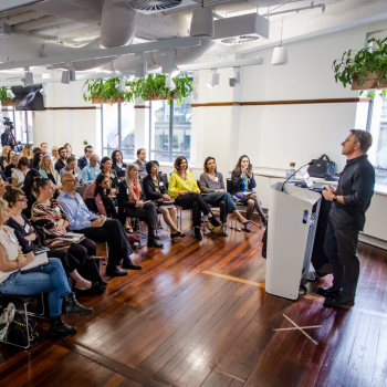 A speaker presenting at a podium to a seated audience in a light-filled venue with timber floors and hanging plants